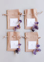 Four beige drawstring pouches with white labels are arranged in a grid, each surrounded by clusters of raw amethyst stones from the Raw Amethyst Crystal Set and other healing crystals on a light background.