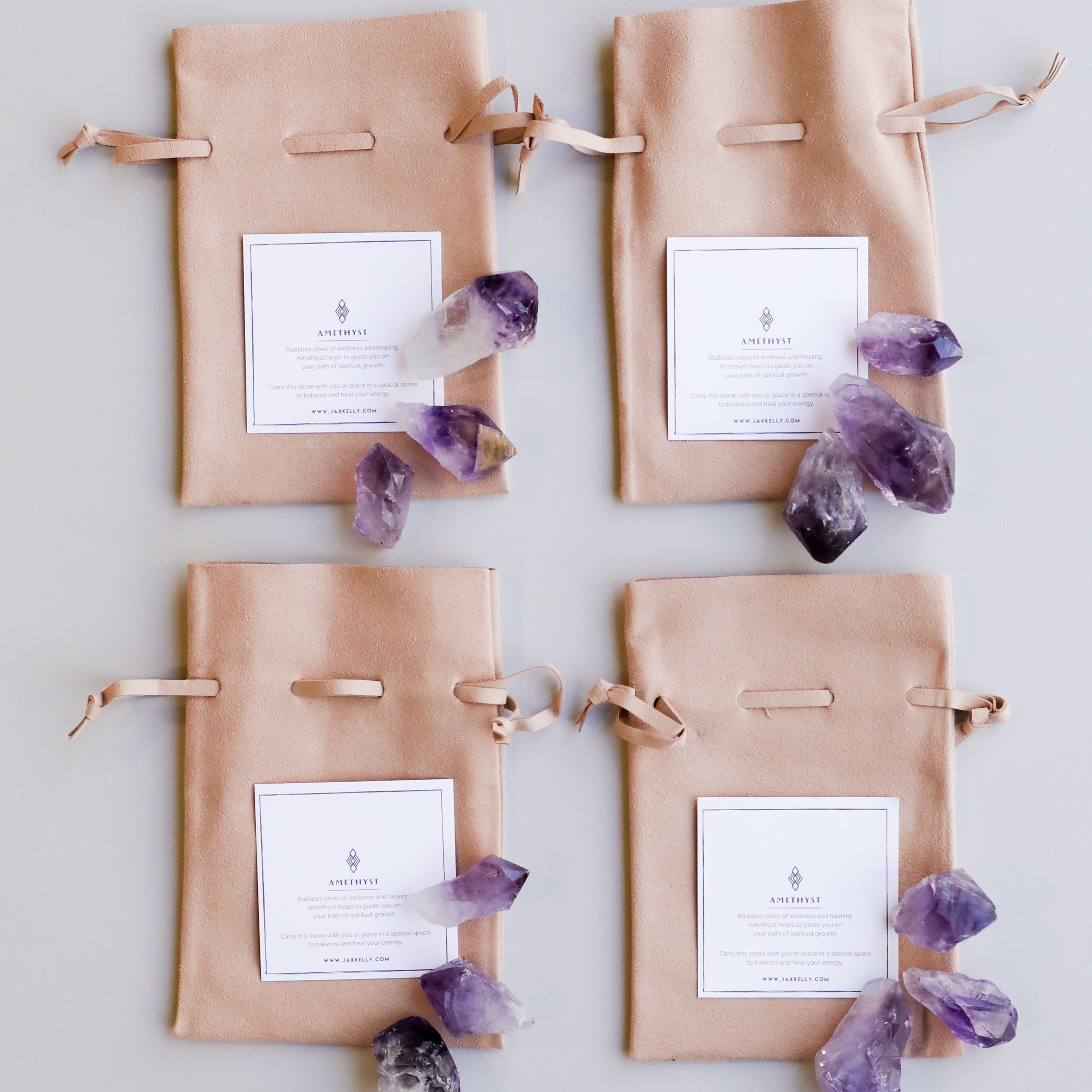 Four beige drawstring pouches with white labels are arranged in a grid, each surrounded by clusters of raw amethyst stones from the Raw Amethyst Crystal Set and other healing crystals on a light background.