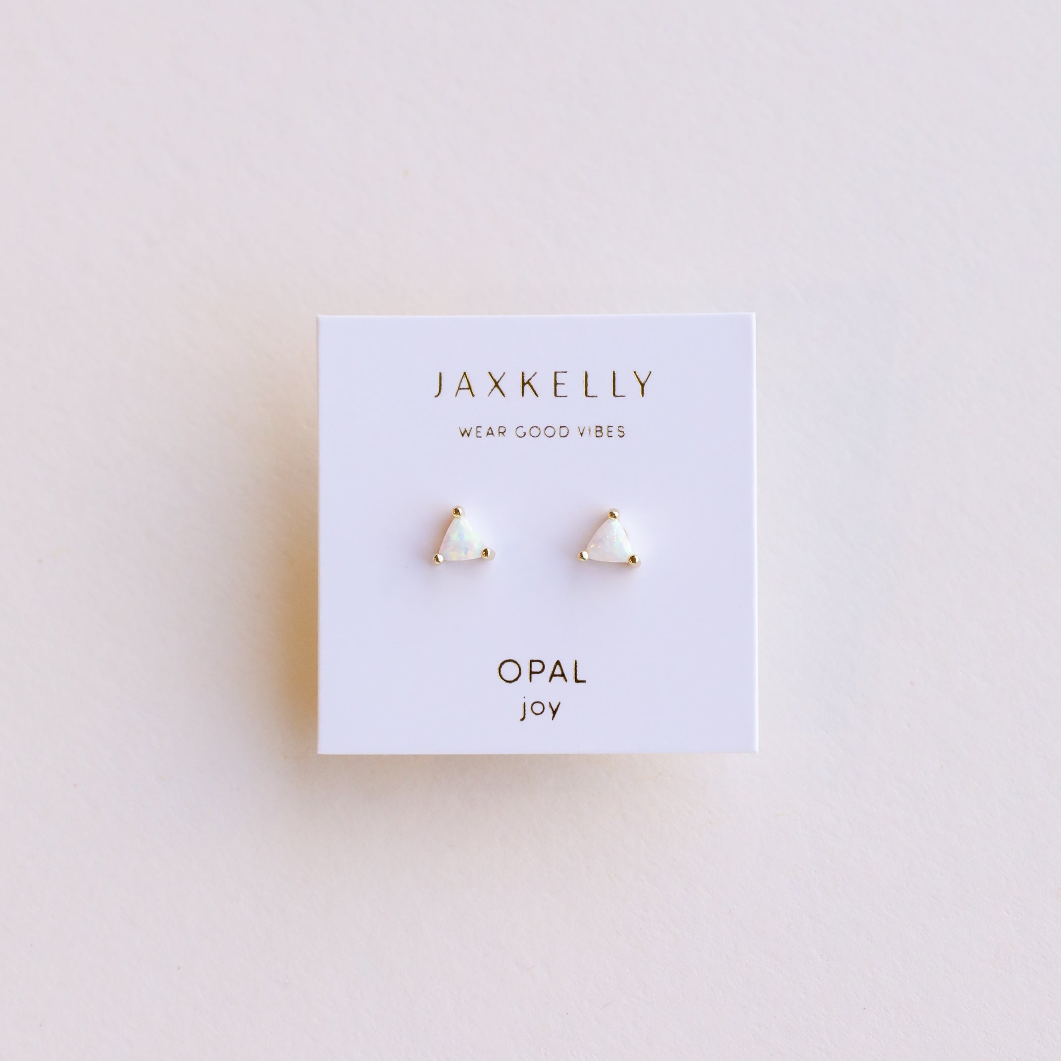 The White Opal Mini Energy Gems - Joy are small, triangular stud earrings with lab-grown white opal and 18k gold plating, shown on a white JaxKelly card that reads WEAR GOOD VIBES and OPAL joy, set against a light, neutral background.