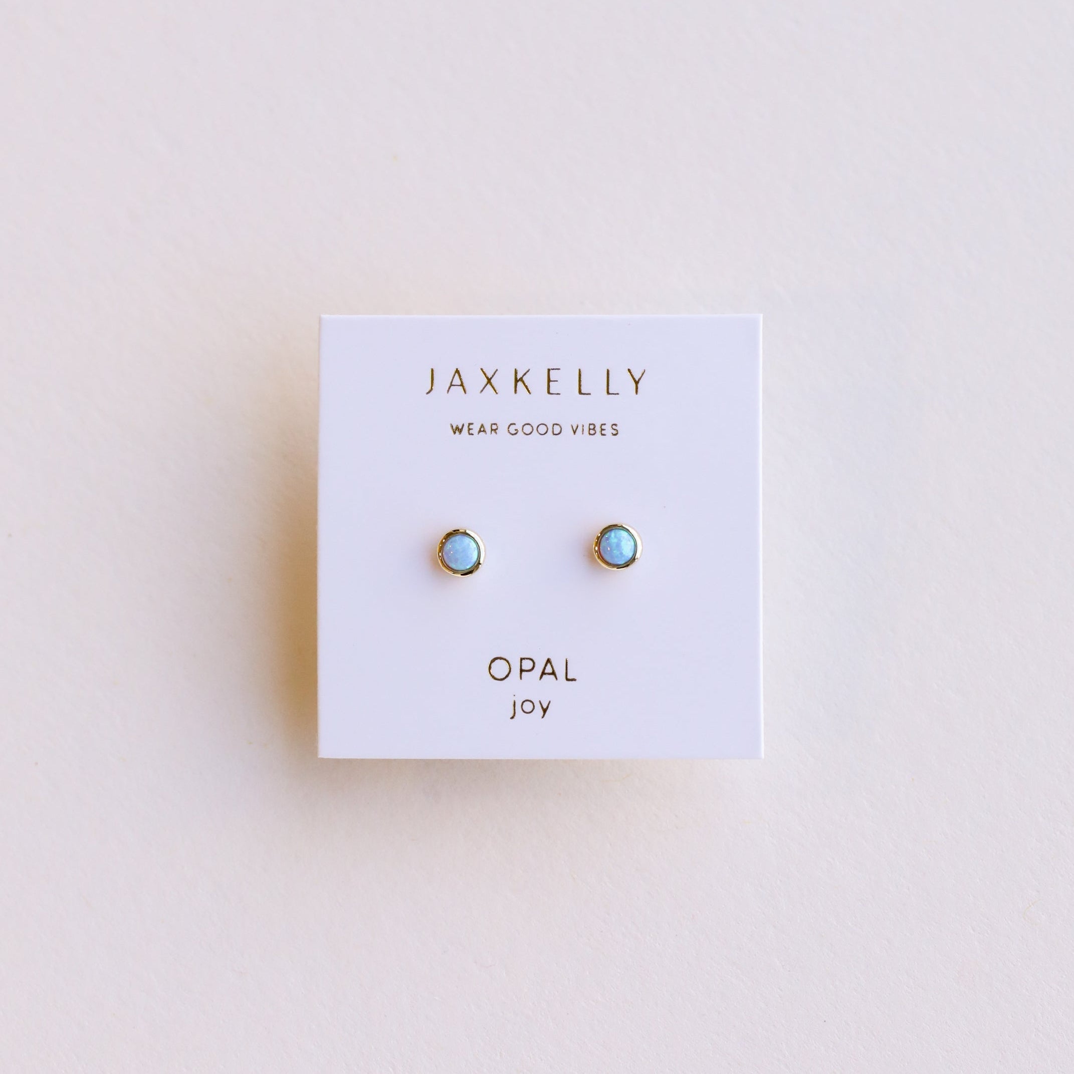 A pair of Simple Stud Earring - Fire Opal, featuring small round lab-grown fire opals in 18k gold plated settings, displayed on a white JaxKelly card labeled "OPAL joy" against a light, neutral background.