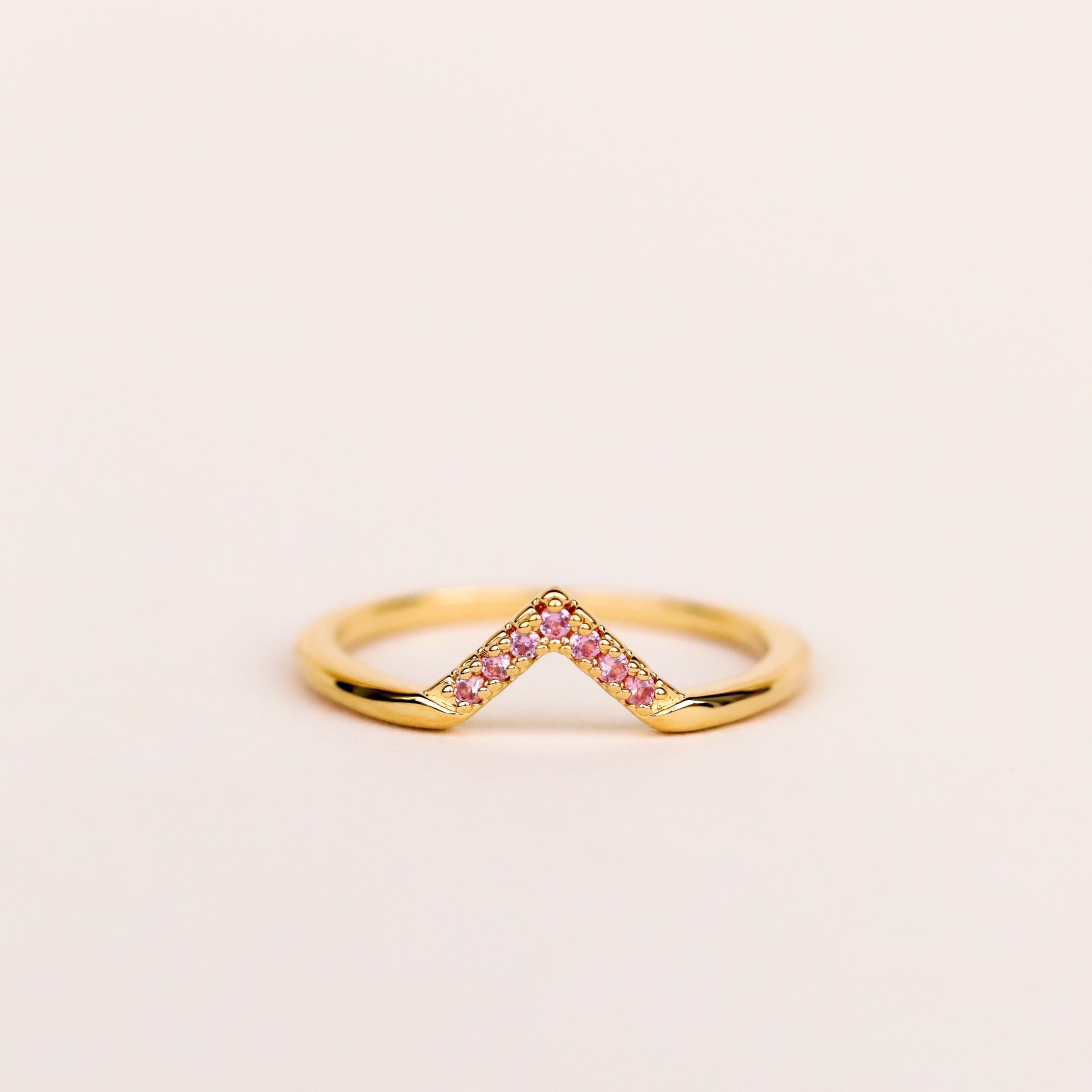 The Pink Lambda Ring features a v-shaped design in 18k gold plating, accented with pink cubic zirconia along the angled edge, shown on a simple light background.
