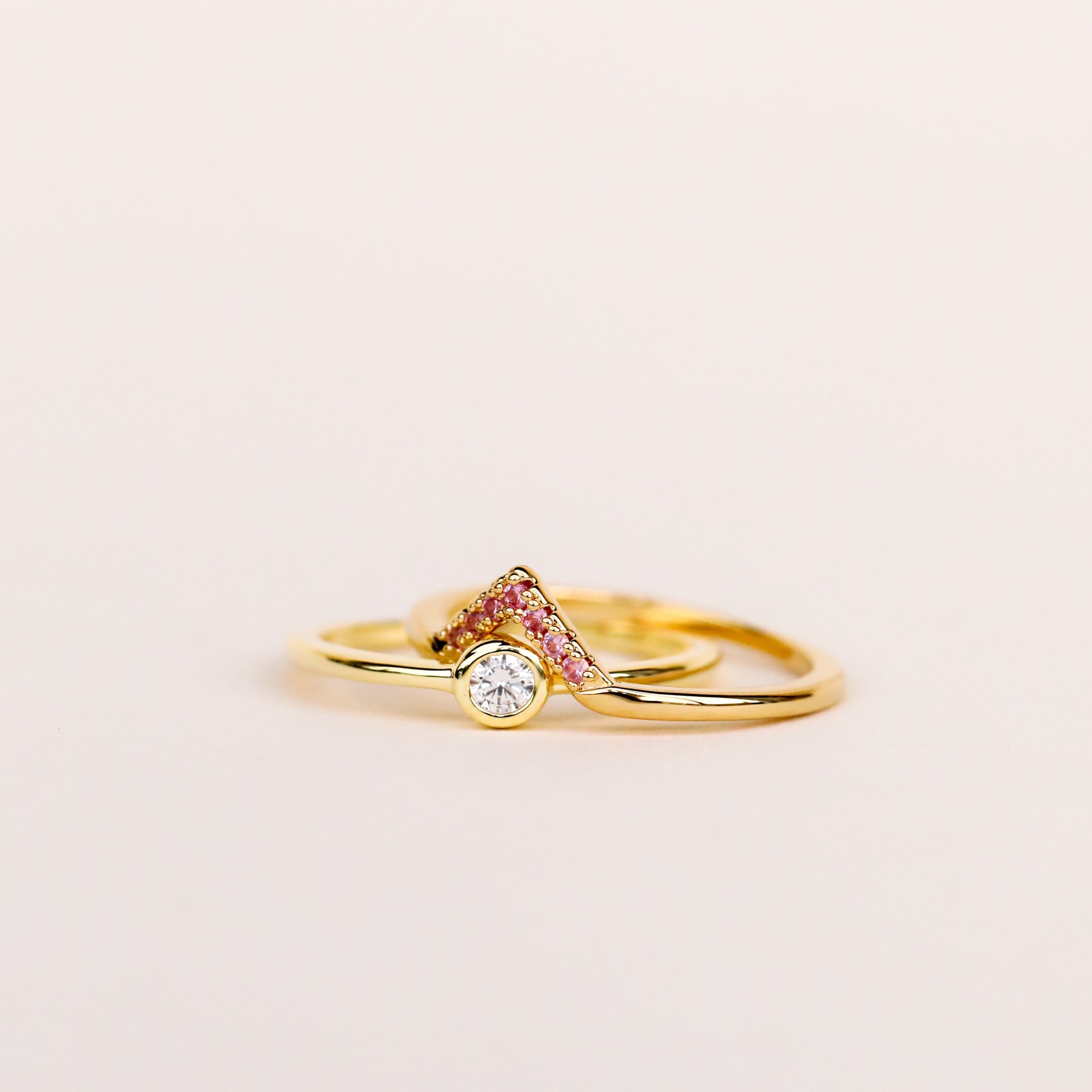 The Pink Lambda Ring features a central round white gemstone on an 18k gold-plated band with Lambda v-shaped accents set with small pink cubic zirconia, shown against a plain light background.