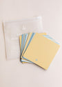 A set of square yellow Note To Self Shower Affirmations™ cards with blue edges and a small leaf icon is spread out beside a translucent white zippered pouch featuring white text.