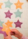 A person holds a purple star-shaped card against a tiled wall decorated with Kids Shower Affirmations™—colorful cards for kids featuring uplifting messages like “I am grateful” and “I dream big and shoot for the stars.”.