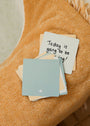 A stack of Note To Self Shower Affirmations™ cards sits on orange textured fabric. The top card reads "Today is going to be amazing!" in black handwritten text; the blue card beneath features a small white flower icon.