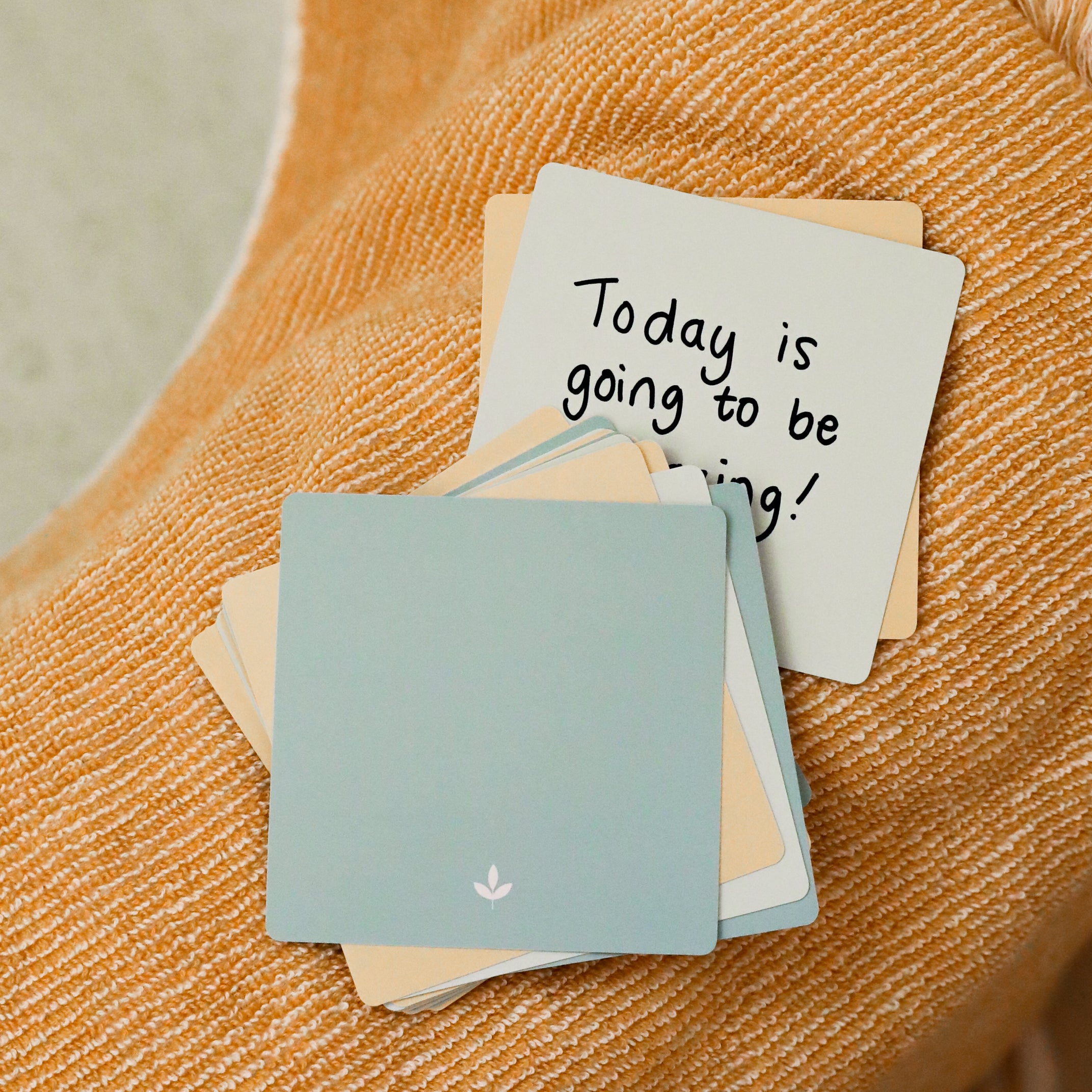 A stack of Note To Self Shower Affirmations™ cards sits on orange textured fabric. The top card reads "Today is going to be amazing!" in black handwritten text; the blue card beneath features a small white flower icon.