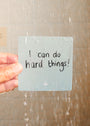A hand holds a Note To Self Shower Affirmations™ card under running water. The waterproof card, covered in droplets, reads “I can do hard things!” with background tiles visible.