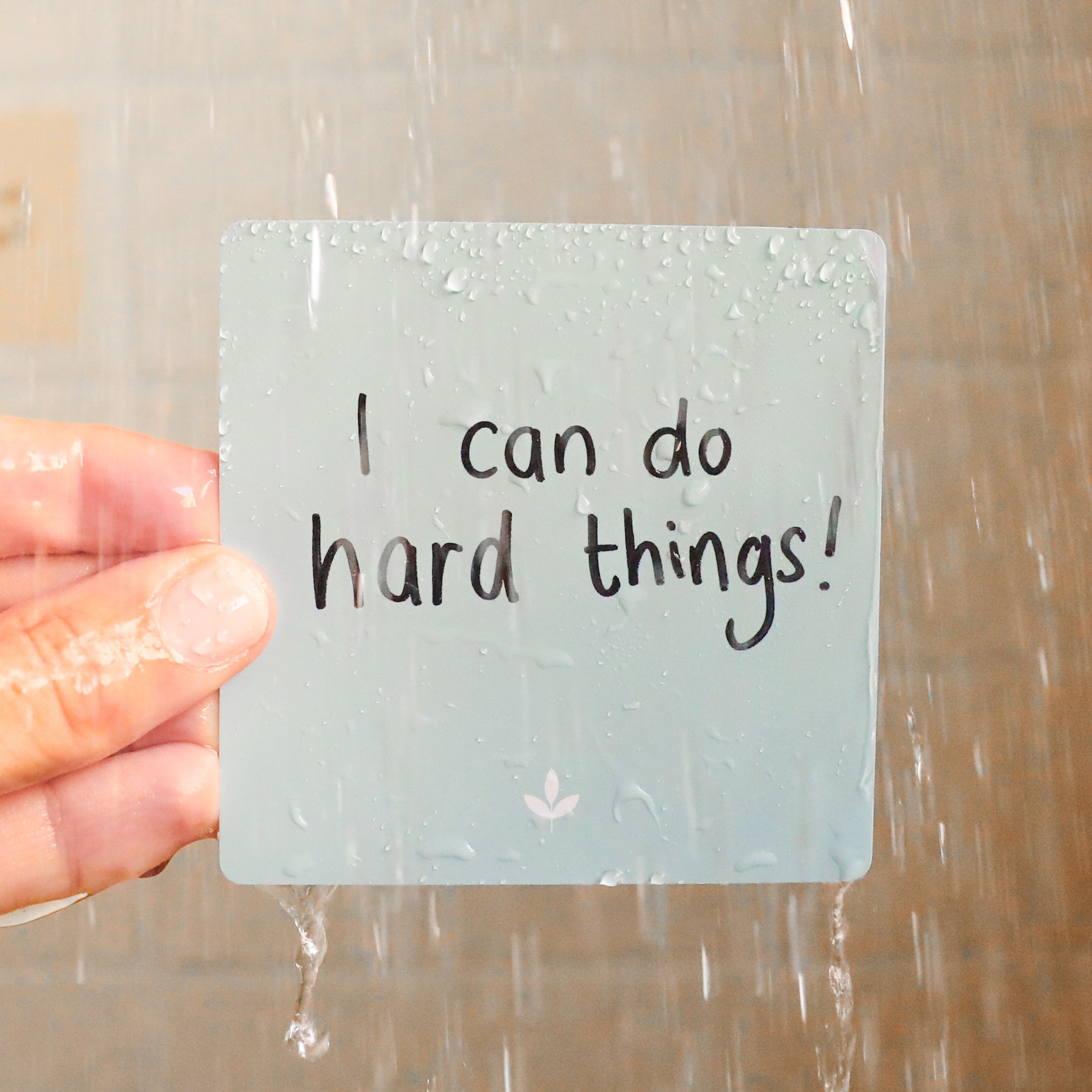 A hand holds a Note To Self Shower Affirmations™ card under running water. The waterproof card, covered in droplets, reads “I can do hard things!” with background tiles visible.