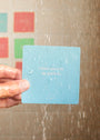 A hand holds a blue Teens Shower Affirmations™ waterproof card with water droplets in the shower. The card reads, "I don’t have to be perfect." Blurred colorful tiles are visible in the background—ideal for teens.