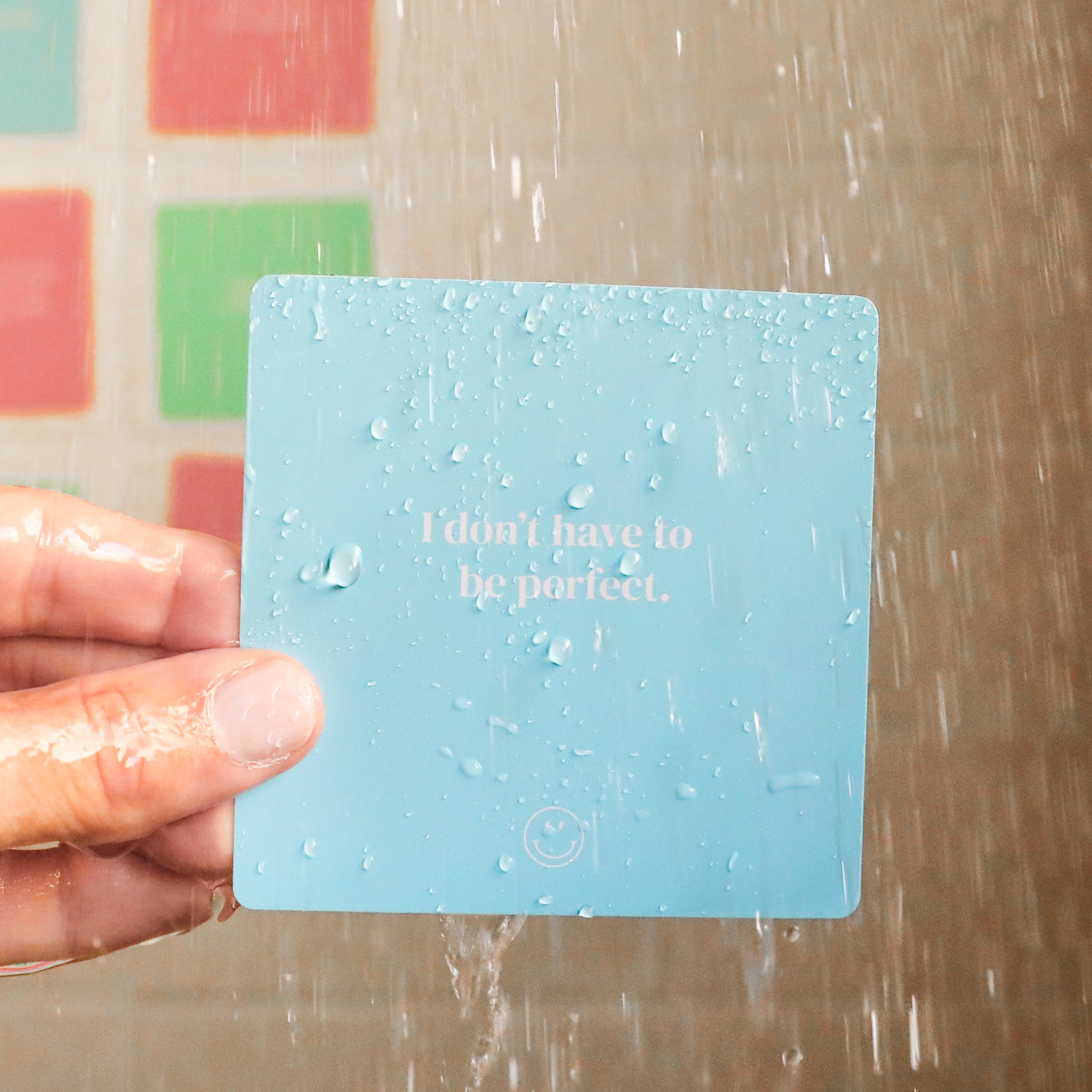 A hand holds a blue Teens Shower Affirmations™ waterproof card with water droplets in the shower. The card reads, "I don’t have to be perfect." Blurred colorful tiles are visible in the background—ideal for teens.