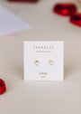 A pair of gold plated stud earrings with lab-grown white opal hearts, labeled Tiny White Opal Heart by JaxKelly, displayed on a white card against a neutral background with blurred red accents around the edges.