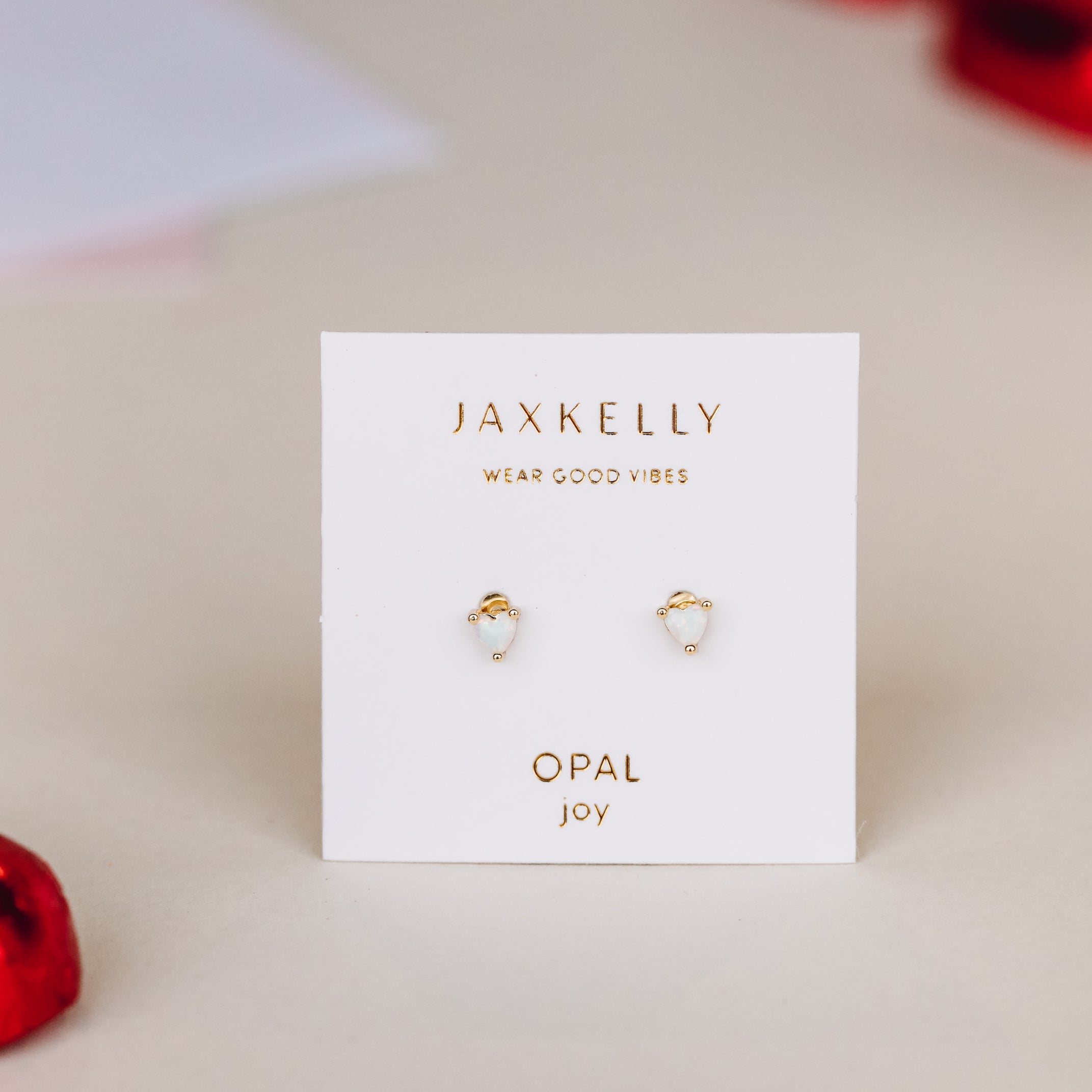 A pair of gold plated stud earrings with lab-grown white opal hearts, labeled Tiny White Opal Heart by JaxKelly, displayed on a white card against a neutral background with blurred red accents around the edges.