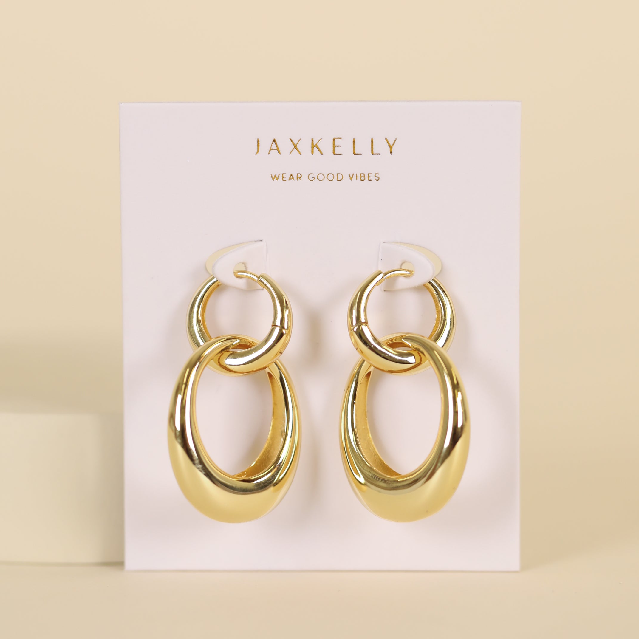 The Coupled Hoop - Earring features a shiny double oval hoop design with 18k gold plating, displayed on a white JaxKelly Wear Good Vibes card against a beige background.