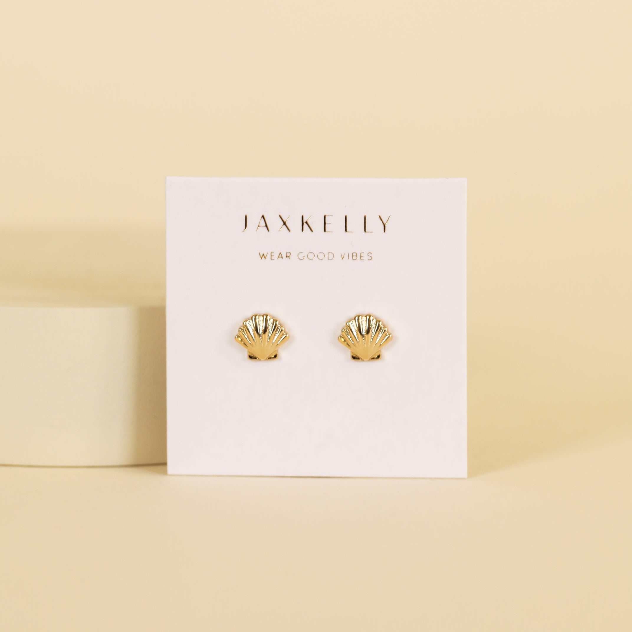 A pair of hypoallergenic Seashell Stud earrings in gold, shaped like shells, are displayed on a white JaxKelly card with the brand slogan “Wear Good Vibes” printed above the earrings.