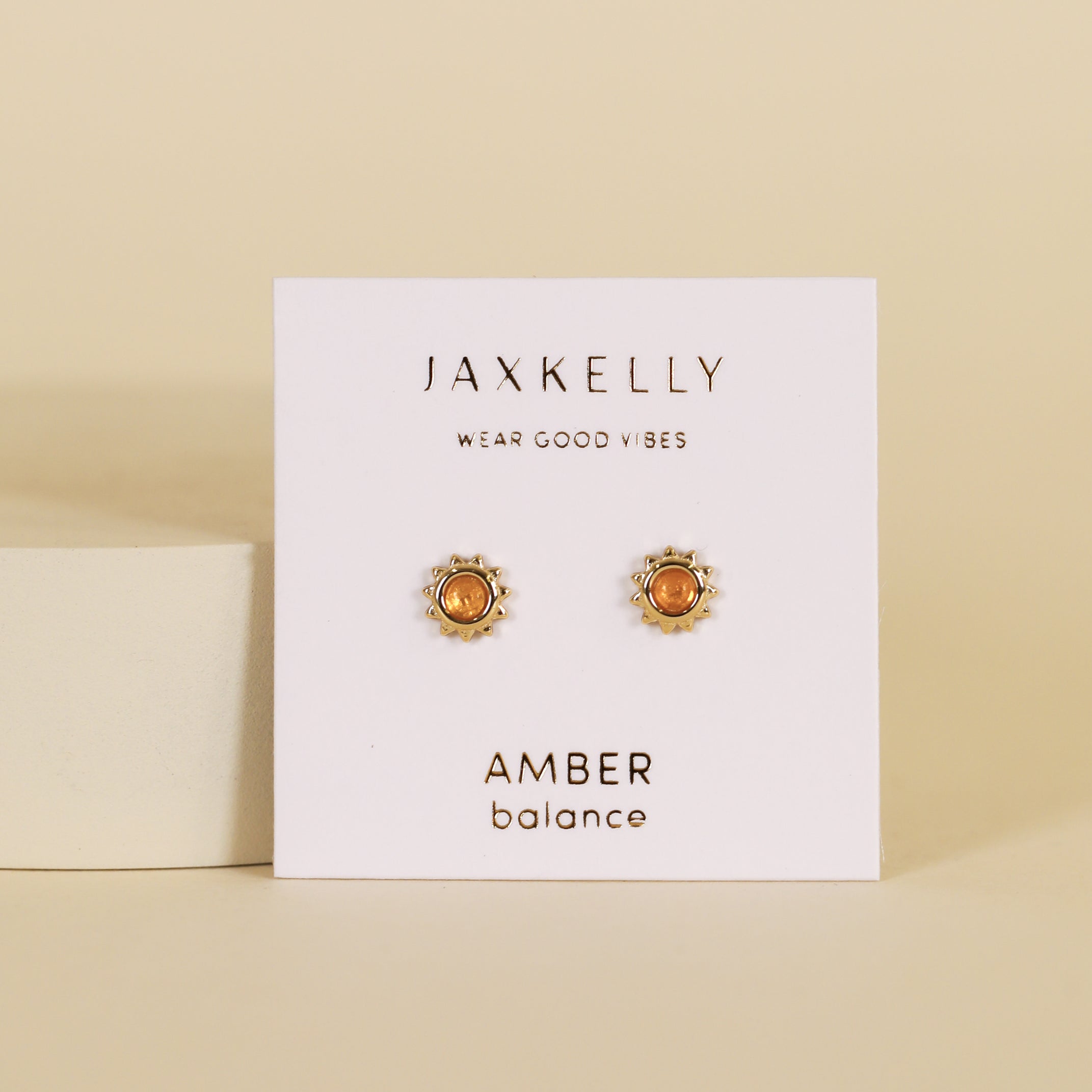 Amber Sun Stud: Gold plated earrings with amber stones displayed on a white card labeled “JaxKelly Wear Good Vibes” and “Amber Balance” against a beige background.