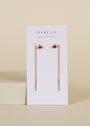 A pair of JaxKelly Studded Threader - Pink CZ earrings, tarnish-free gold chain drops with a small pink stone and vertical gold bar, shown on a white JaxKelly card with "wear good vibes" tagline against a beige background.