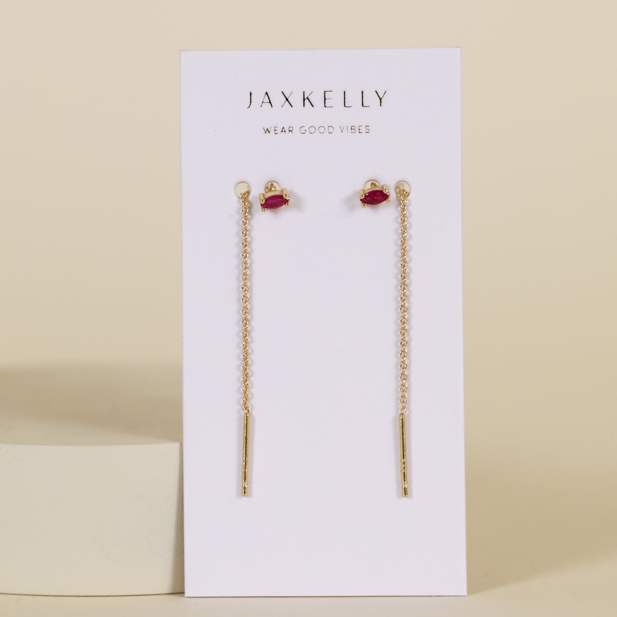 A pair of JaxKelly Studded Threader - Pink CZ earrings, tarnish-free gold chain drops with a small pink stone and vertical gold bar, shown on a white JaxKelly card with "wear good vibes" tagline against a beige background.