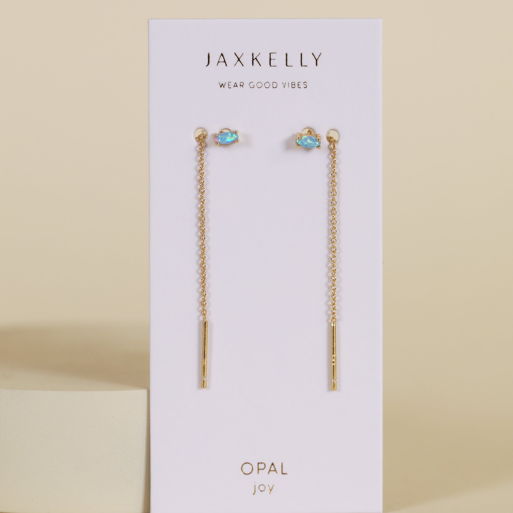 The JaxKelly Studded Threader - Fire Opal earrings, featuring 18K gold plating and lab-grown fire opal studs, are shown on a white card labeled JaxKelly and Opal Joy against a neutral background.