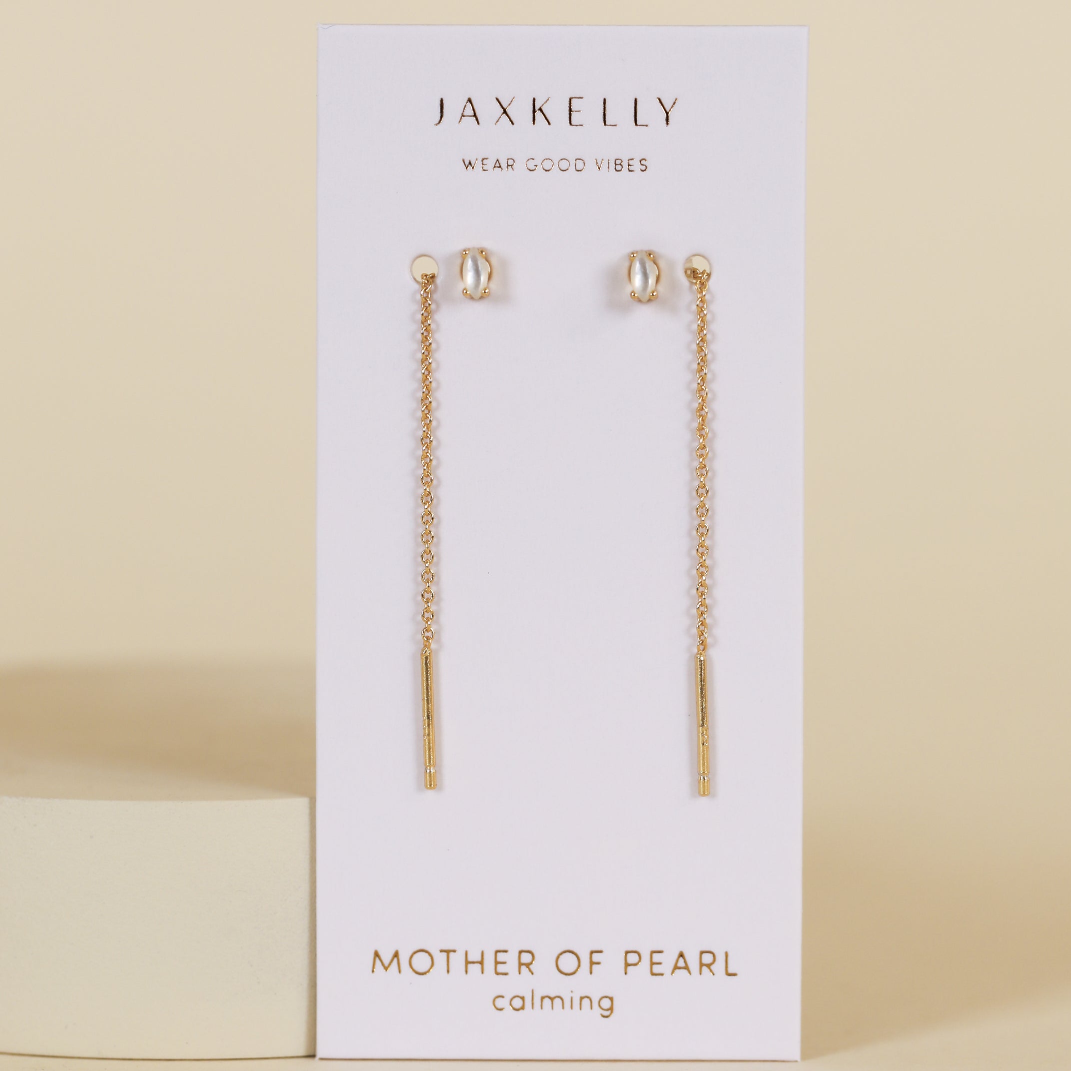 The JaxKelly Studded Threader - Mother of Pearl earrings, featuring gold chains and pearl studs with mother-of-pearl accents, are shown on a white JaxKelly card against a neutral beige background.