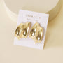 A pair of Gold Bold Wave sculptural statement studs by JaxKelly, featuring 18k gold plating, displayed on a white JaxKelly card reading "WEAR GOOD VIBES," set on a cream-colored surface.