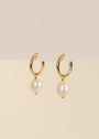 The Pearl Drop Earring features gold-plated hoops, round white freshwater pearl pendants, and sterling silver posts, shown against a cream-colored background.