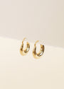 The Sculptural Wide Hoop by JaxKelly features a bold, thick rounded design in shiny 18K gold plating, displayed on the edge of a white surface with a light beige background.