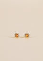 The Amber Sun Stud earrings are small gold studs featuring a round amber-colored center, shown on a plain beige background.