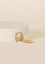 The Triple Arch Hoop - Earring is a pair of small gold hoops with layered detailing, featuring triple arch designs—one displayed upright on a white circular platform, the other lying flat on a beige surface.