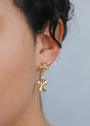 Close-up of a person wearing Double Daisy Drop Earrings—gold-plated earrings featuring two daisy shapes, one on the earlobe and a larger floral drop below. The neutral background is softly blurred.