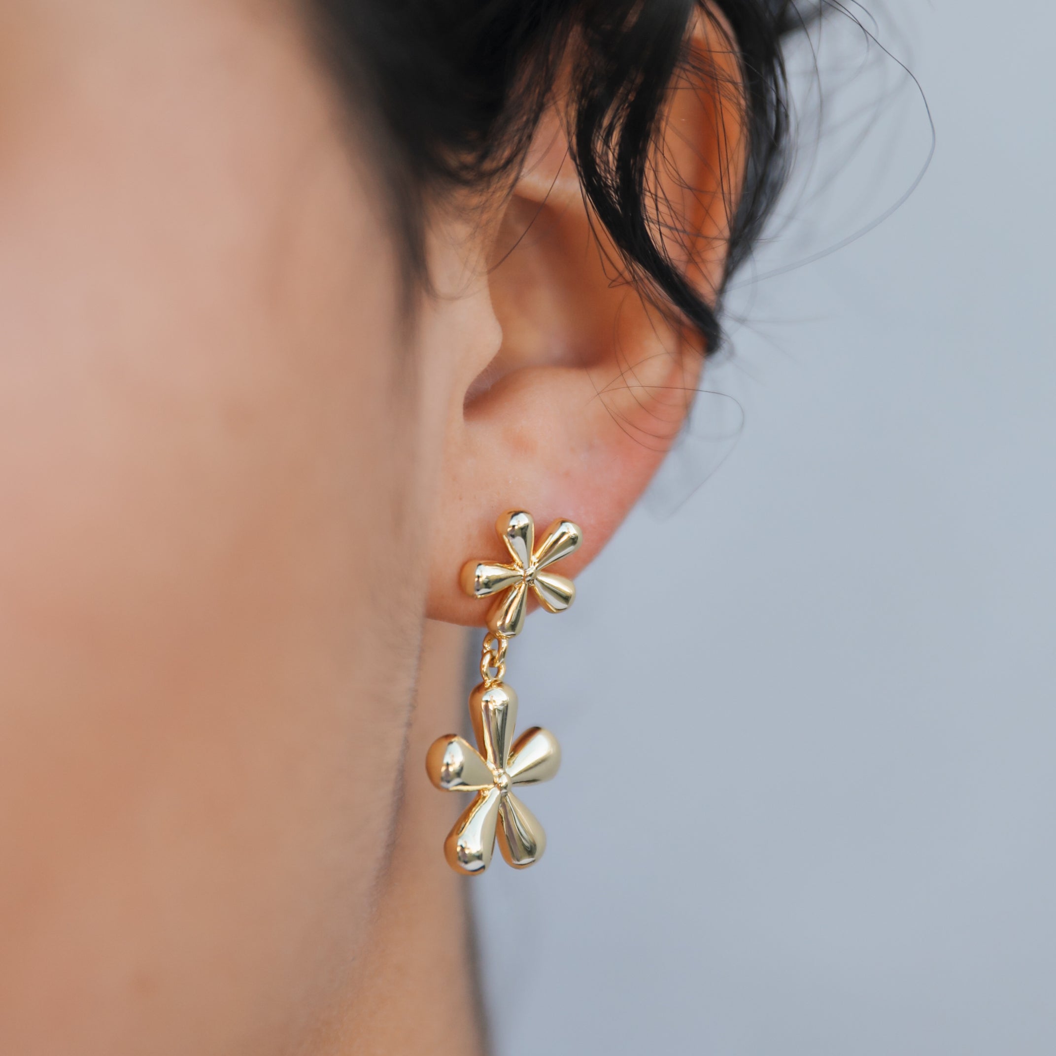 Close-up of a person wearing Double Daisy Drop Earrings—gold-plated earrings featuring two daisy shapes, one on the earlobe and a larger floral drop below. The neutral background is softly blurred.