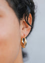 Close-up of a person's ear with short, curly dark hair, wearing JaxKelly's Sculptural Wide Hoop earrings in shiny, tarnish-free 18K gold plating against a neutral background.