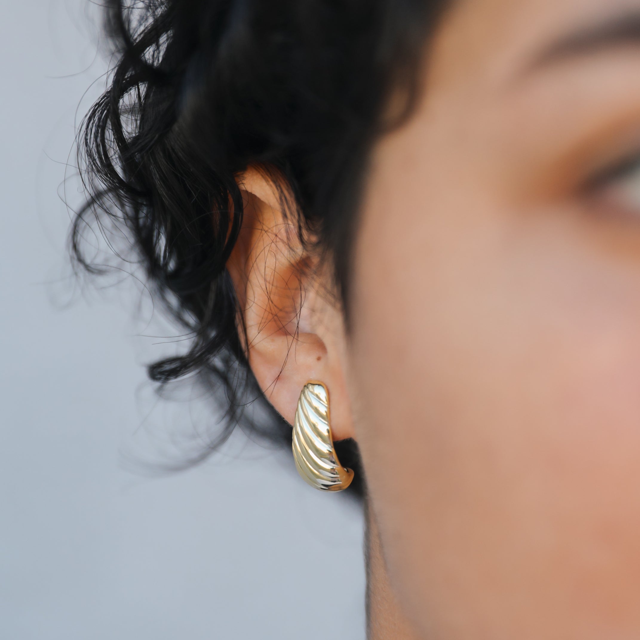 Close-up of a person with short curly dark hair wearing the Bold Swirl - Earring, a textured gold plated hoop, in their left ear, with focus on this statement jewelry against a neutral background.