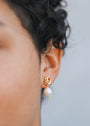 Close-up of a person wearing the Pearl Knot - Earring, featuring 18k gold-plated knot detail and a single freshwater pearl drop. The blurred background enhances the earring's shine against curly hair.