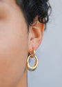 Close-up of a person’s ear wearing the Coupled Hoop - Earring with 18k gold plating. The lightweight double hoops rest against smooth skin, with dark curly hair nearby and a softly blurred background for a gentle touch.