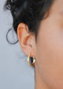 Close-up of a person's ear wearing the Textured Hoop - Earring, a small gold hoop with a leaf-like design and timeless hoops style. Short, curly hair is partially visible against a softly blurred background.