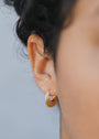 Close-up of a person's ear wearing the Triple Arch Hoop - Earring, featuring small gold-plated triple arches with sterling silver posts. The dark, curly hair and blurred background accentuate the earring's elegant design.