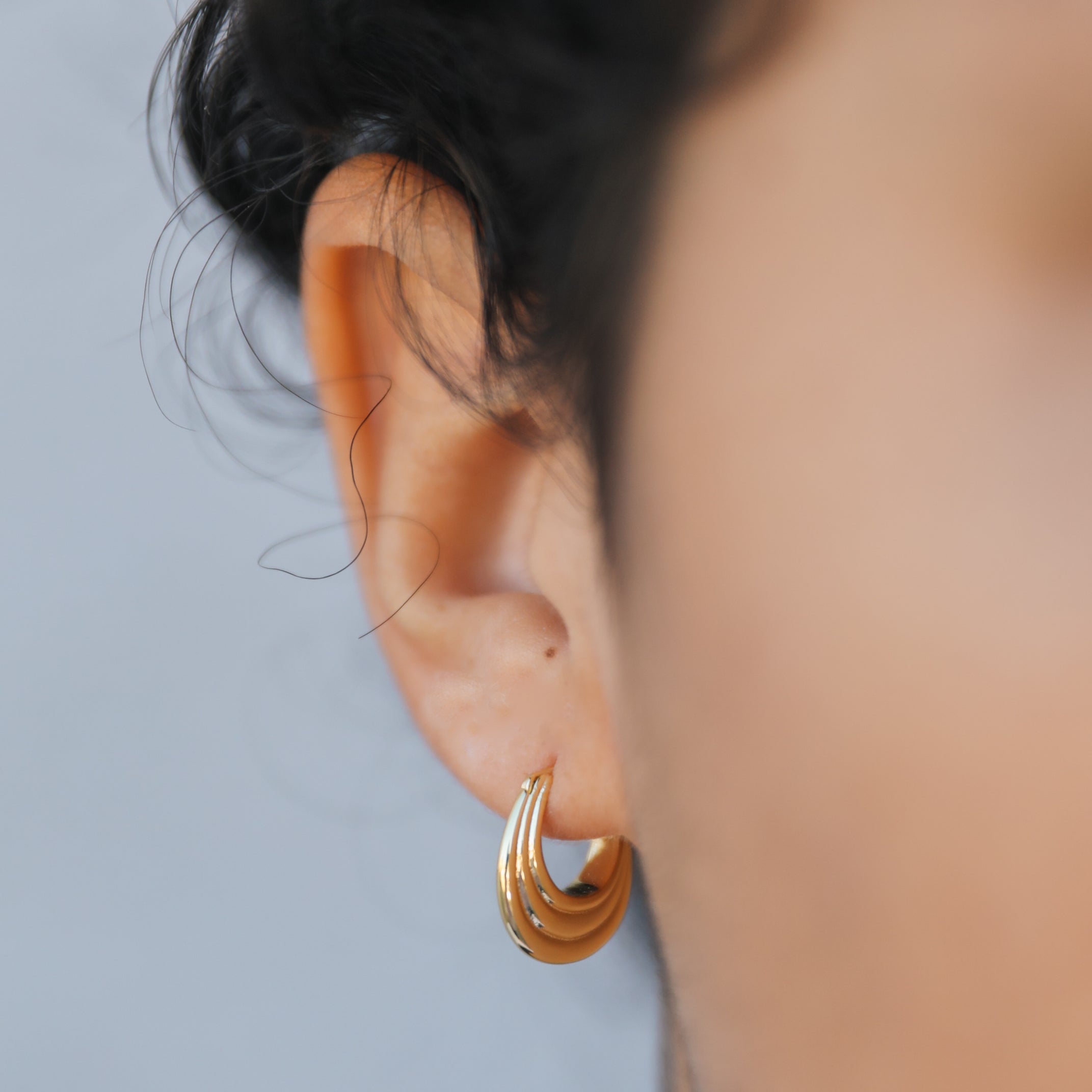 Close-up of a person's ear wearing the Triple Arch Hoop - Earring, featuring small gold-plated triple arches with sterling silver posts. The dark, curly hair and blurred background accentuate the earring's elegant design.