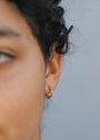 Close-up of a person's ear wearing the Double Hoop - Earring, a small gold plated hoop, with part of their curly hair and blurred facial features visible against a plain background.