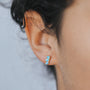 Close-up of a person wearing the Offset Trio - Fire Opal: a small gold plated stud earring featuring multiple light blue stones in a curved line. The person's dark, curly hair is partially visible in the background.