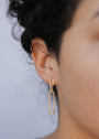 Close-up of a person's ear with short, curly dark hair, wearing the Pave Link Drop - Earring featuring two elongated chain links and 18k gold plating. The plain background is softly blurred.