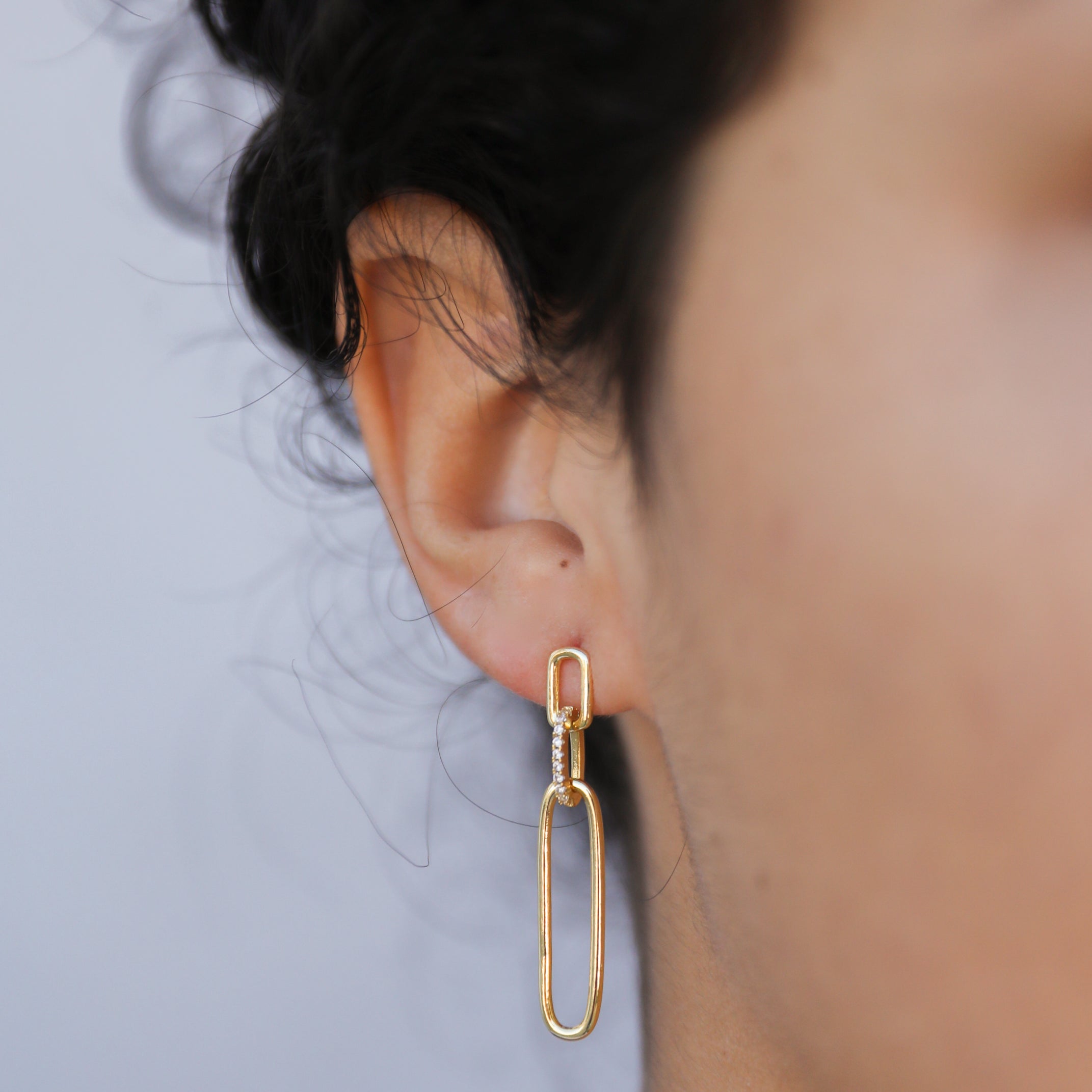 Close-up of a person's ear with short, curly dark hair, wearing the Pave Link Drop - Earring featuring two elongated chain links and 18k gold plating. The plain background is softly blurred.
