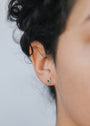 A close-up of a person's ear with curly dark hair, wearing the Malachite Sun Stud—featuring a malachite stone encircled by 18k gold plating. The background is blurred, and part of the person’s face is visible.