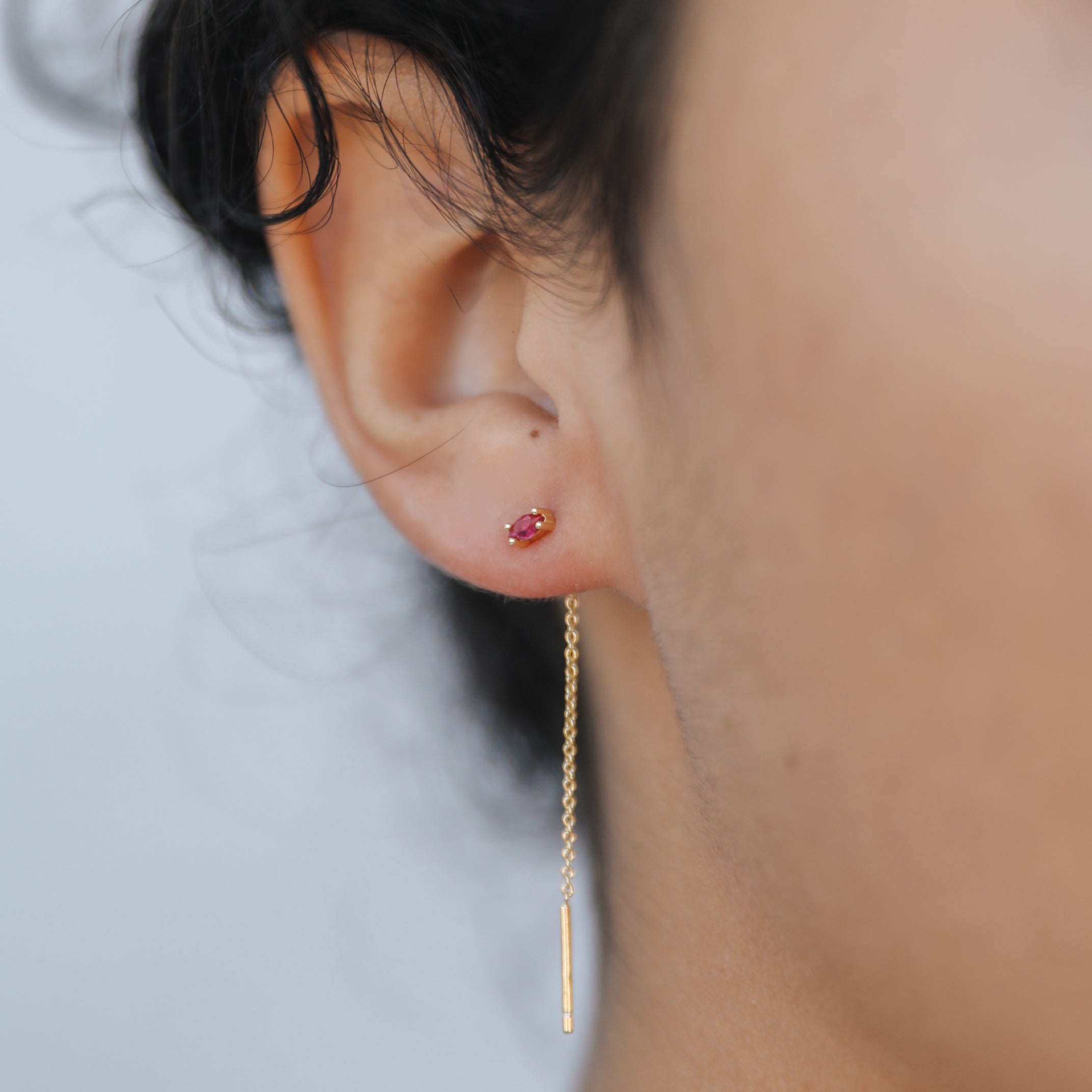Close-up of a person’s ear with dark curly hair, wearing JaxKelly's Studded Threader - Pink CZ earring. The small pink stud and long gold threader stand out against the plain, blurred background, showcasing tarnish-free elegance.