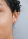 Close-up of a person’s ear wearing JaxKelly’s Studded Threader - Mother of Pearl earring, featuring a small stud and a long, thin gold chain. The background is plain, highlighting the skin and hair in soft focus.