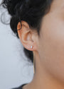 Close-up of a person's ear wearing JaxKelly's Studded Threader – White Opal, featuring a lab-grown white opal stud in the upper lobe and an 18K gold plated threader in the lower lobe. Plain background highlights their dark, curly hair.