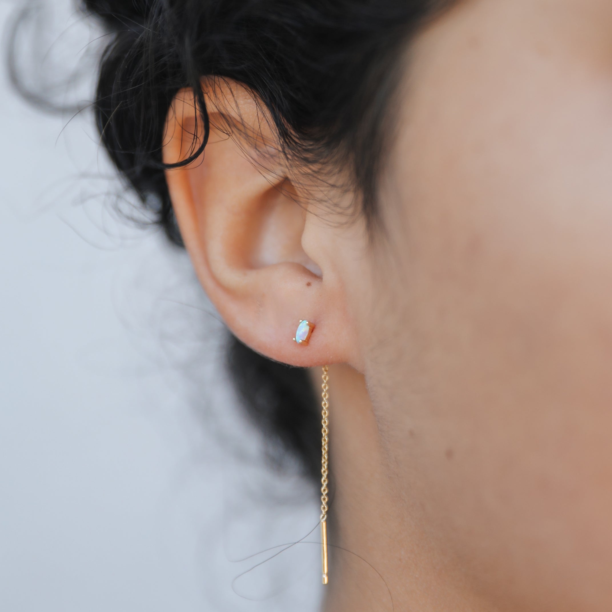 Close-up of a person's ear wearing JaxKelly's Studded Threader – White Opal, featuring a lab-grown white opal stud in the upper lobe and an 18K gold plated threader in the lower lobe. Plain background highlights their dark, curly hair.