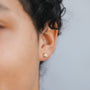 Close-up of a dark, curly-haired person wearing the Seashell Stud - Earring—a small, gold, hypoallergenic piece—against a plain light gray background.