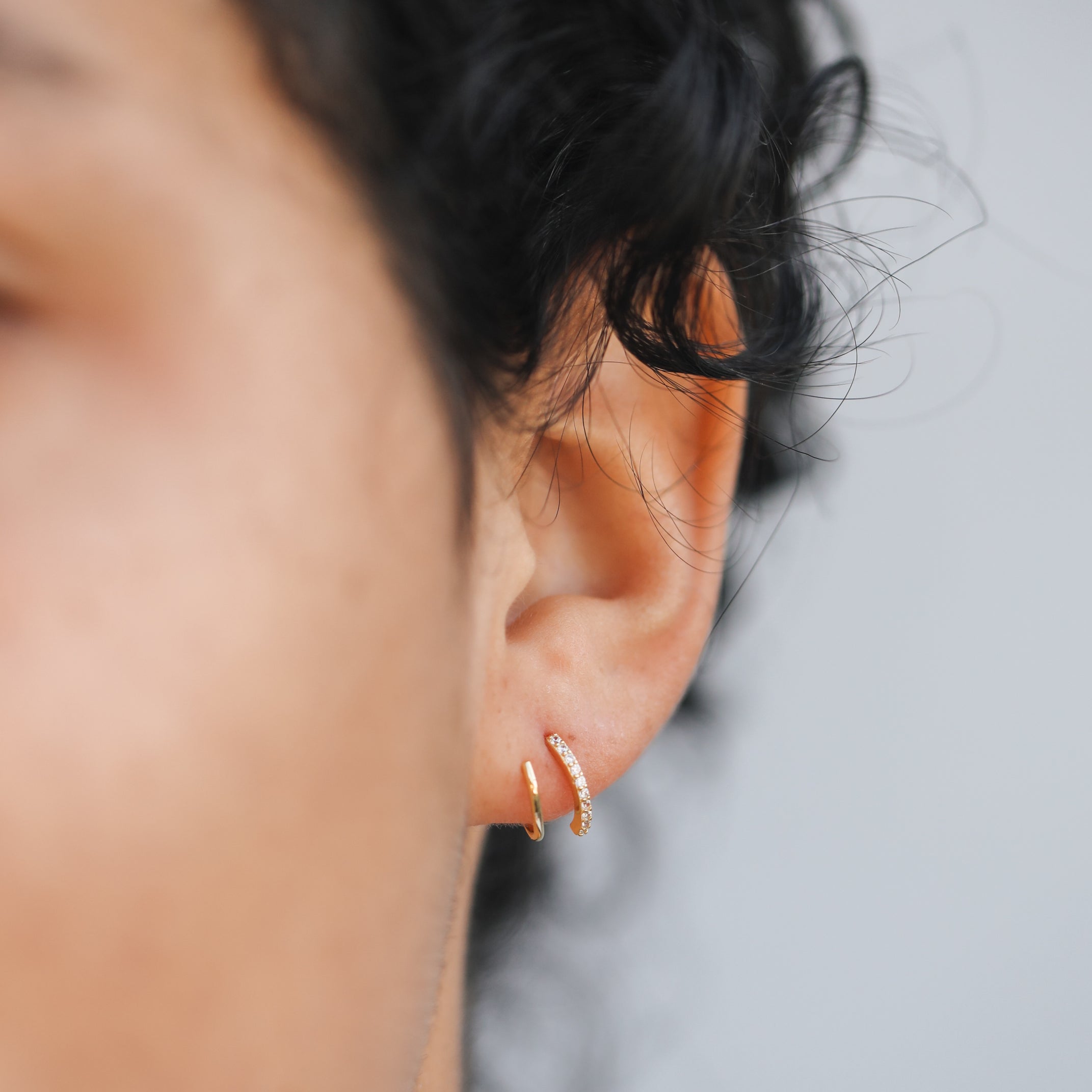 A close-up of a person's ear shows two gold hoops—one smooth, the other the sparkling Pave Spiral - Earring—with curly dark hair and part of their face softly visible in the background.