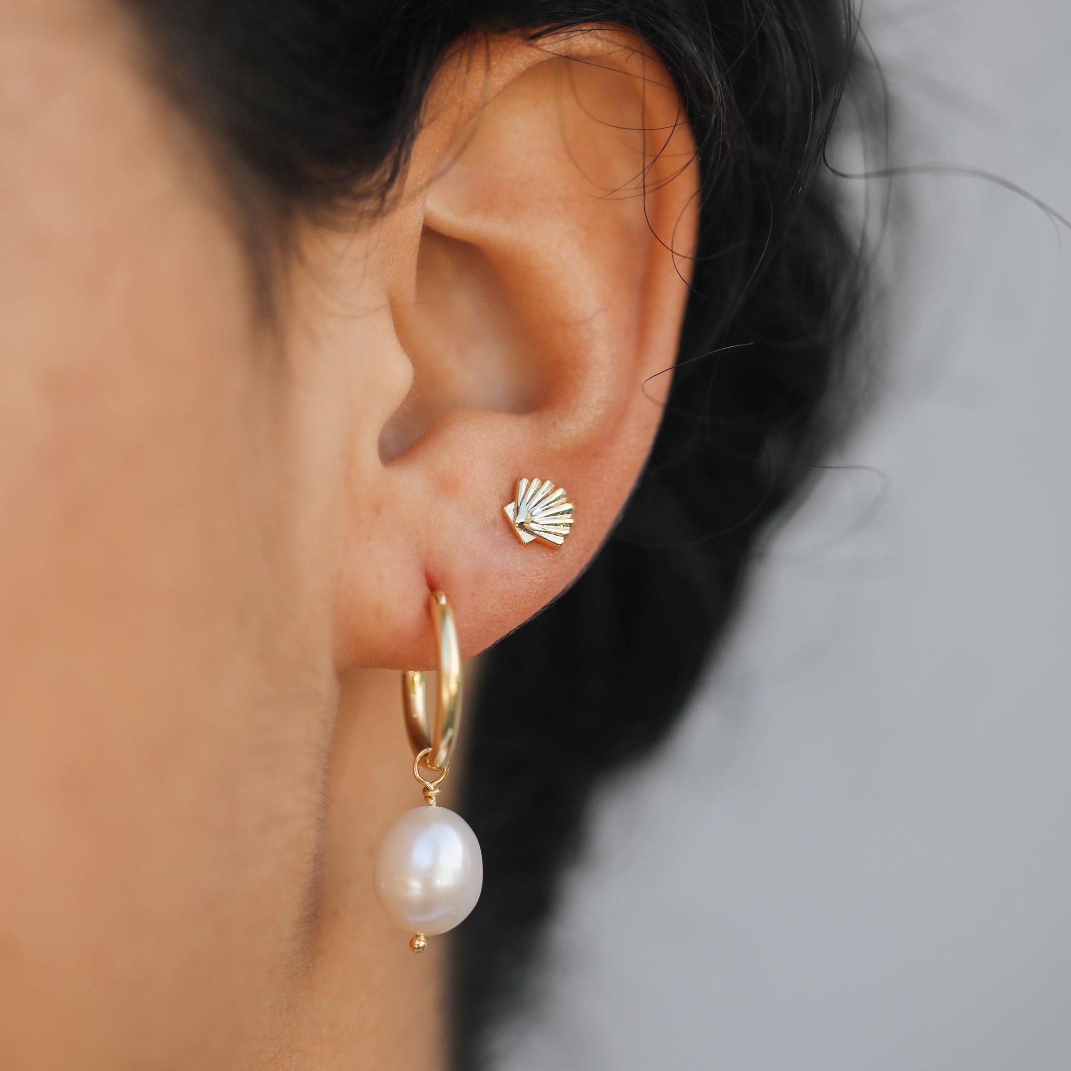 A close-up of a person’s ear wearing two earrings: a small gold seashell stud in the upper lobe and the Pearl Drop Earring—a gold plated hoop with a hanging freshwater pearl—in the lower lobe, set against a soft neutral background.