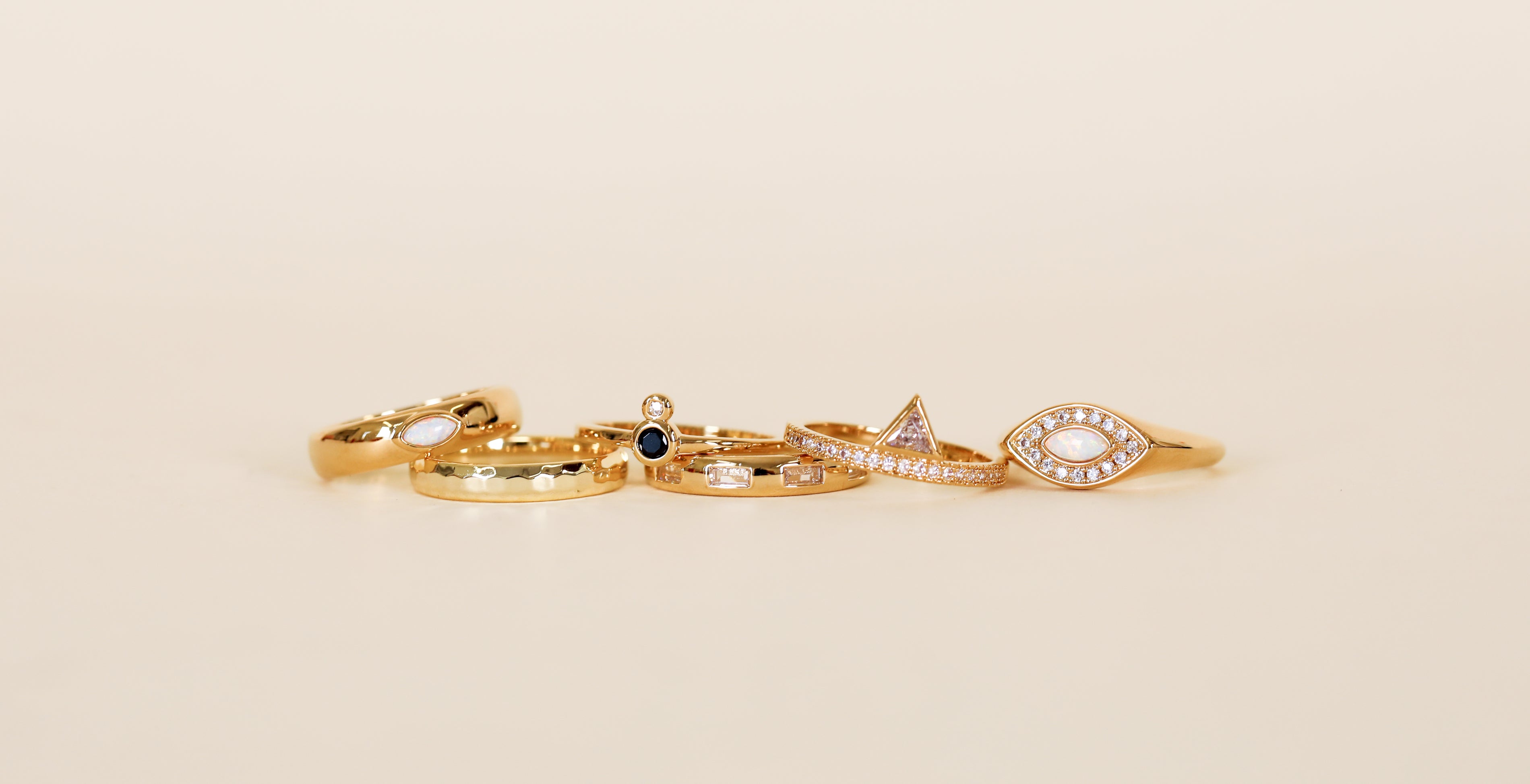 Five Mystery Rings in assorted gold designs are arranged on a plain beige background, featuring gemstones, a geometric triangle, and an eye-shaped motif with small embedded diamonds—ideal as a special jewelry gift or for a ring sale.